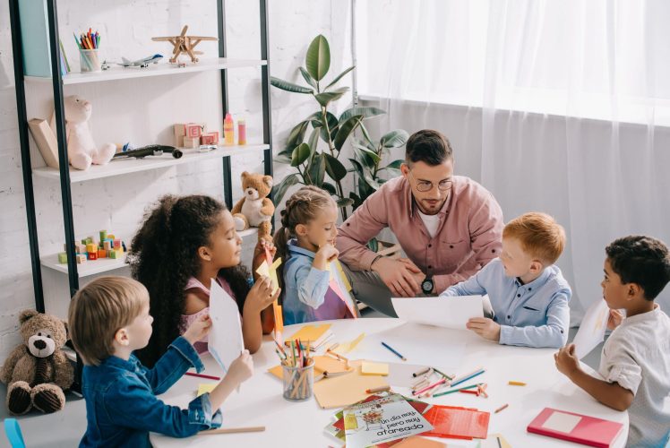 teacher and interracial preschoolers at table with paints and papers in classroom